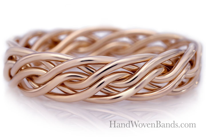 A close-up of an intricately handwoven rose gold band with a shiny finish, displayed on a reflective white surface.