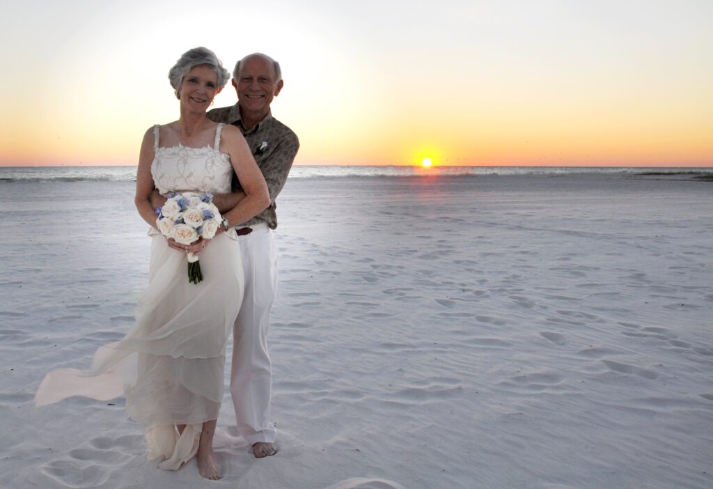 An older couple in wedding attire smiling on a beach at sunset, with the woman holding a bouquet and showcasing a wedding ring.