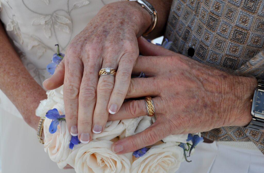 Close-up of an elderly couple's hands with wedding rings from a renowned store, placed over a bouquet of white roses.