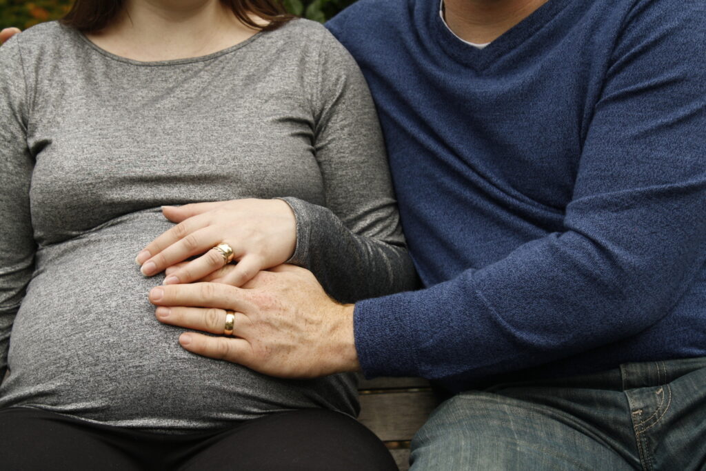 A pregnant woman and a man sitting together, with the man's hand on her belly, both wearing long-sleeved shirts and showcasing wedding rings from nearby stores.