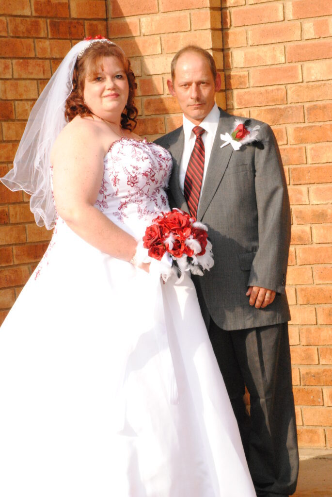 A bride in a white and red wedding dress and a groom in a black suit with a red boutonniere from a wedding ring store standing together against a brick wall.