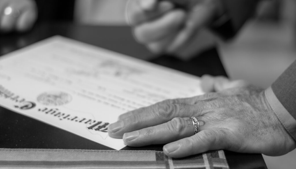 A black and white photo of a person's hand resting on a document, with a wedding ring visible, while another person writes on the document with a pen.