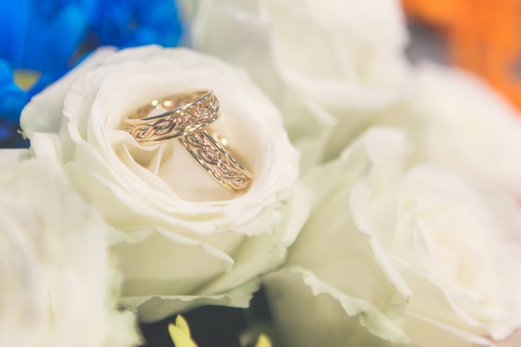 Two gold wedding ring stores offer intricate designs resting on white roses, with a soft-focus background.