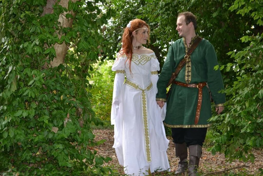 A couple dressed in medieval costumes, walking hand in hand and admiring wedding rings, smiling at each other in a lush green garden.