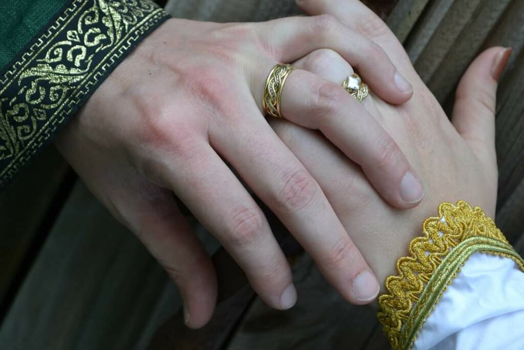Close-up of a hand wearing a wedding ring from one of the premier wedding ring stores, placed atop another hand, both adorned with elegant, embroidered sleeves.