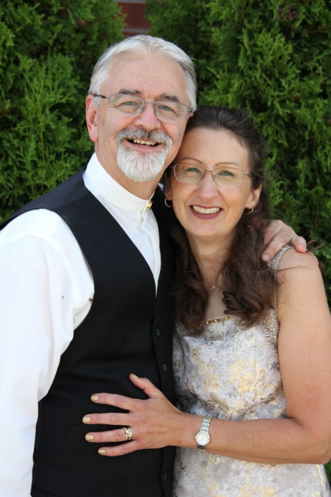 An elderly couple, a man in a black vest and a woman in a white floral dress, smiling and embracing in front of green bushes, beautifully showcasing their wedding rings.