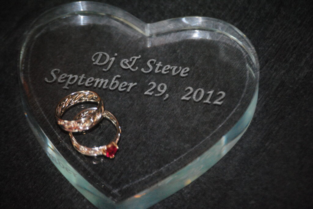 Heart-shaped glass keepsake engraved with "dj & steve september 29, 2012" next to two intertwined wedding rings on a dark background.