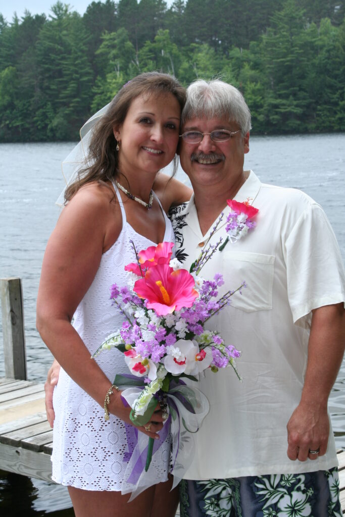 A smiling couple in casual wedding attire holding a bouquet and displaying wedding rings, standing on a dock by a lake.
