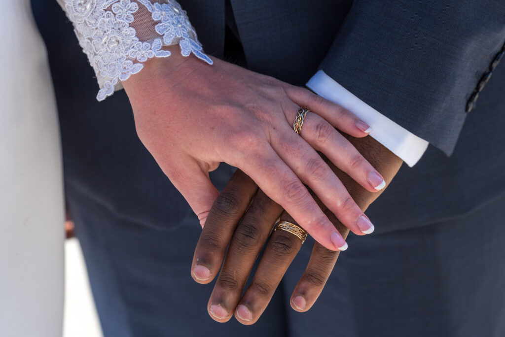 Close-up of a newlywed couple's hands, showcasing their wedding rings from premier wedding ring stores, with the bride in a lace sleeve and the groom in a suit.