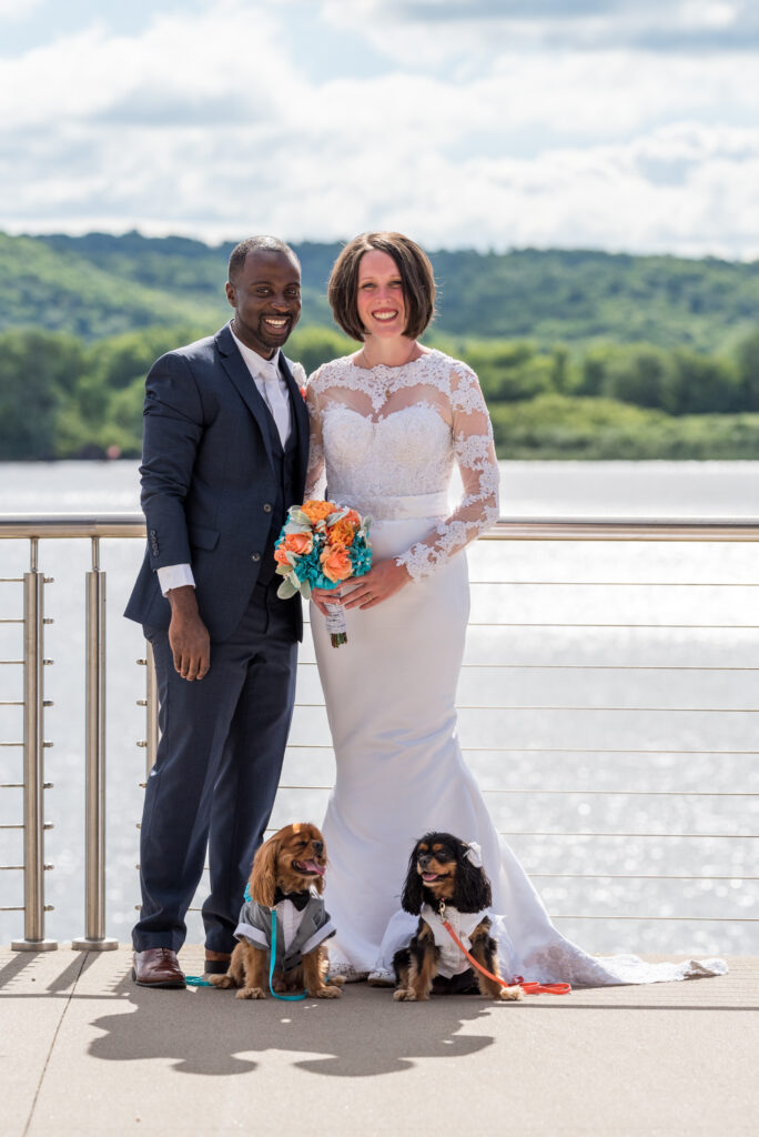 A newlywed couple posing with two King Charles spaniels by a lakeside, the woman in a white lace gown and the man in a navy suit, both showcasing their wedding rings from renowned stores