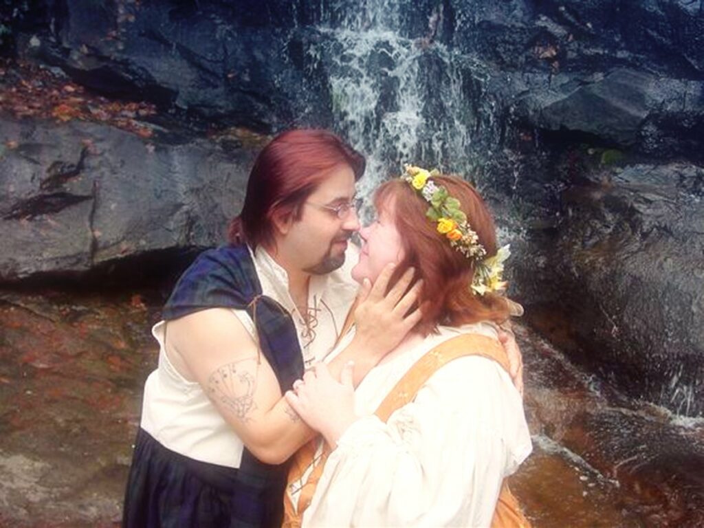 A couple dressed in medieval costumes embraces near a waterfall, exchanging loving gazazes as the man holds the woman's face gently and subtly shows a wedding ring from one of their favorite wedding ring stores.