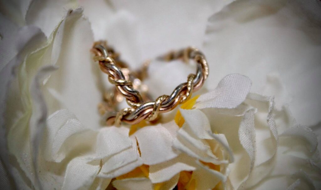 Gold chain bracelet and wedding ring draped over white artificial flowers with yellow centers, highlighted by a soft-focus background.