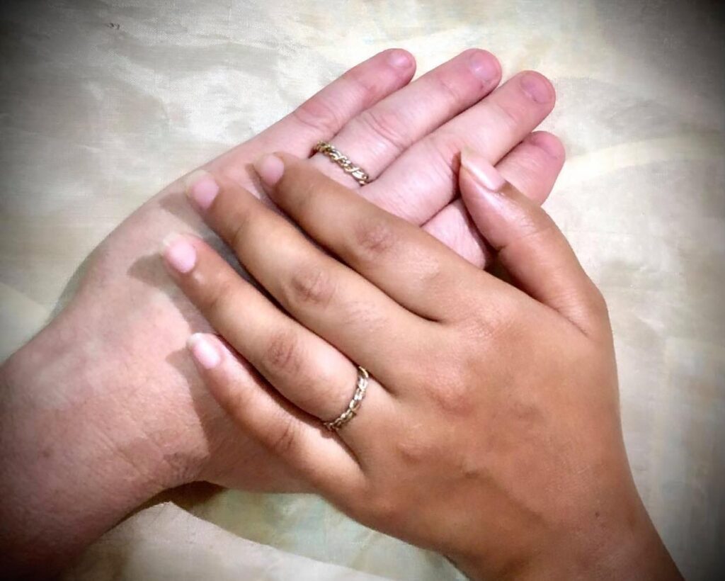 Two hands clasped, one with a wedding ring, on a pale background.