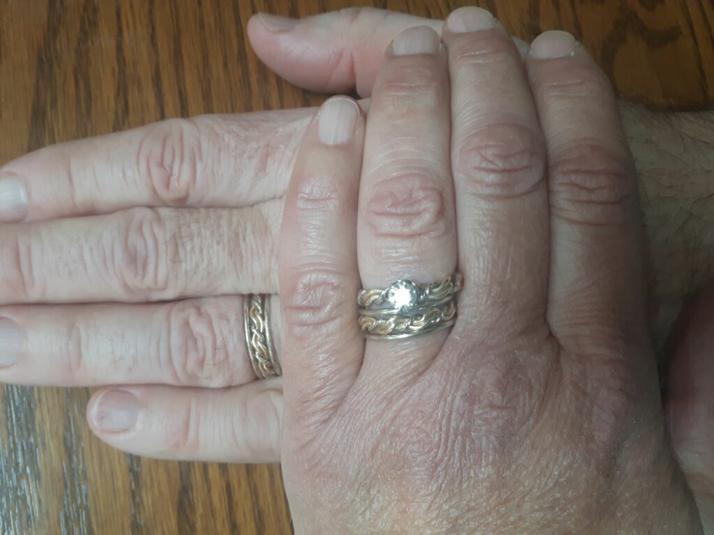 Two hands clasped together on a wooden surface, showing a close-up of their fingers with one wedding ring on each hand.