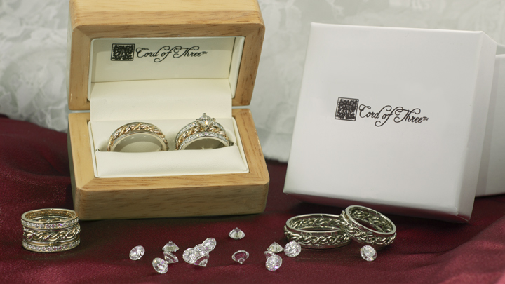 Two ornate Cord of Three Strands wedding rings in a wooden box with the lid open, additional rings and loose gemstones scattered in front, and a white box with logo in the background.