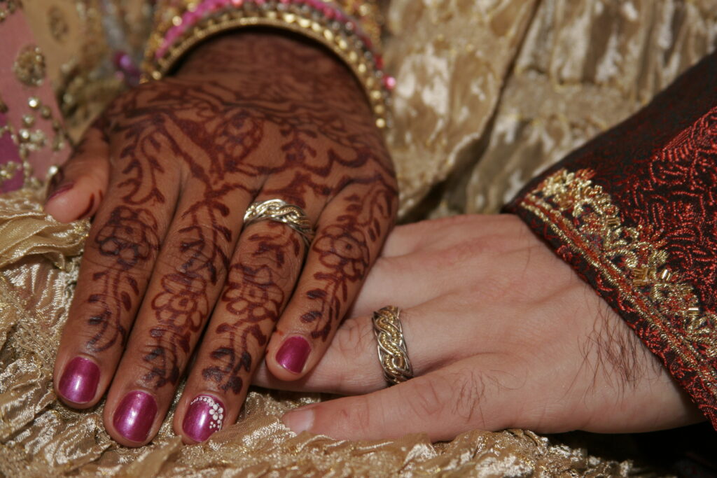 A close-up of a couple's hands, one with elaborately decorated henna designs and the other without, both wearing eight strand double weave wedding rings, resting on a golden fabric.