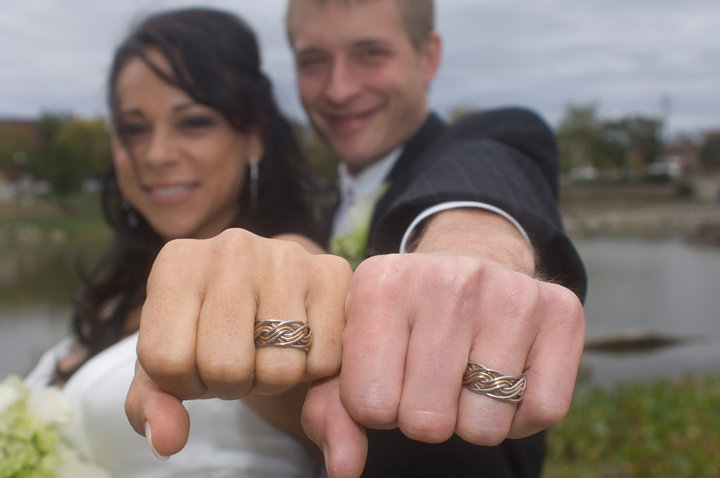 A newlywed couple proudly showing their matching Eight Strand Double Weave silver wedding bands, smiling in the foreground with a soft-focus background.