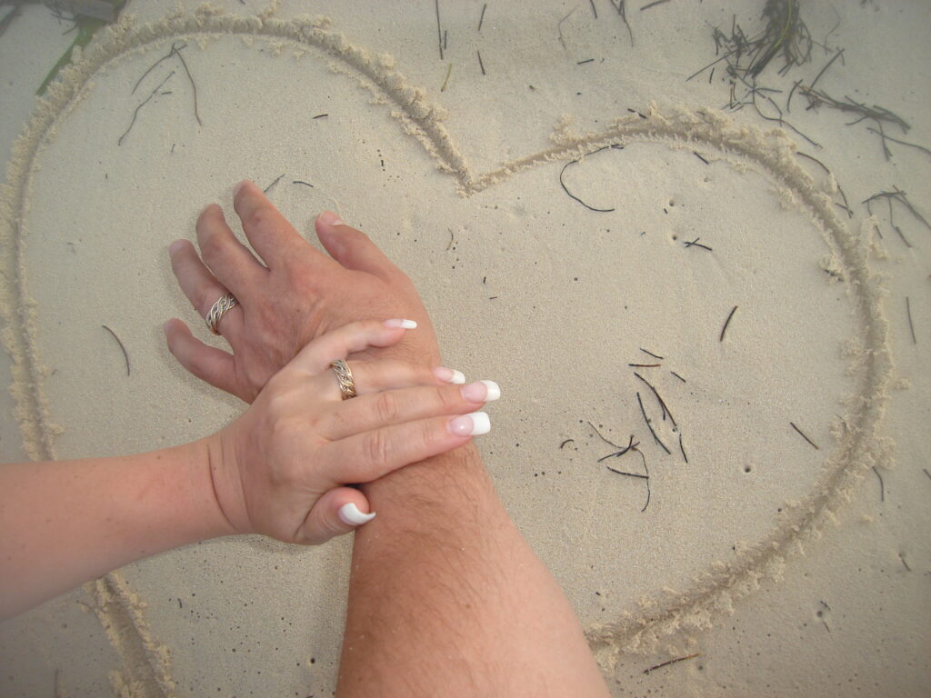 Two hands, one with an Eight Strand Double Weave Ring, clasped together inside a heart shape drawn in the sand.