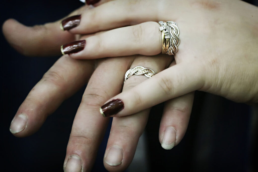 Close-up of two hands clasped together, displaying six strand closed weave silver rings on each hand's ring finger; both sets of fingernails are painted dark red.
