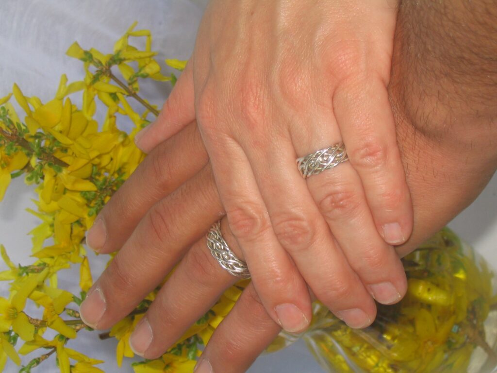 Two hands with eight strand open weave silver rings, one atop the other, resting on a surface with yellow flowers and a glass jar.