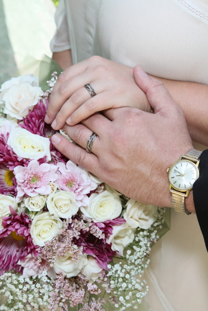 Close-up of a bride and groom's hands with a Seven Strand Ring, over a bouquet of white and pink flowers.