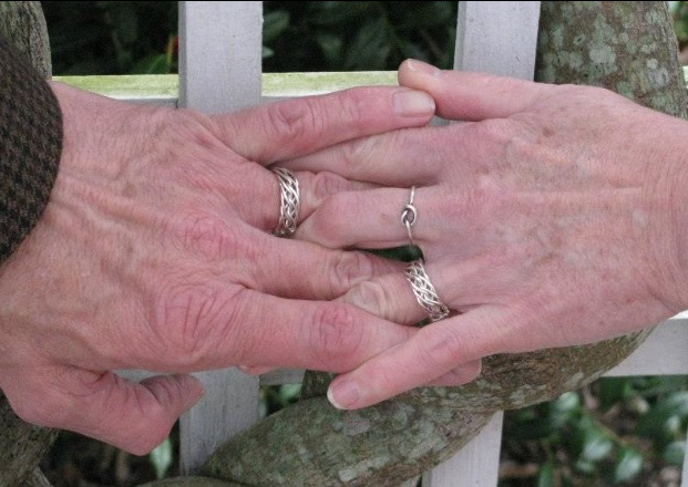 Two elderly hands with an eight strand open weave ring gently touch over a white wooden bench.