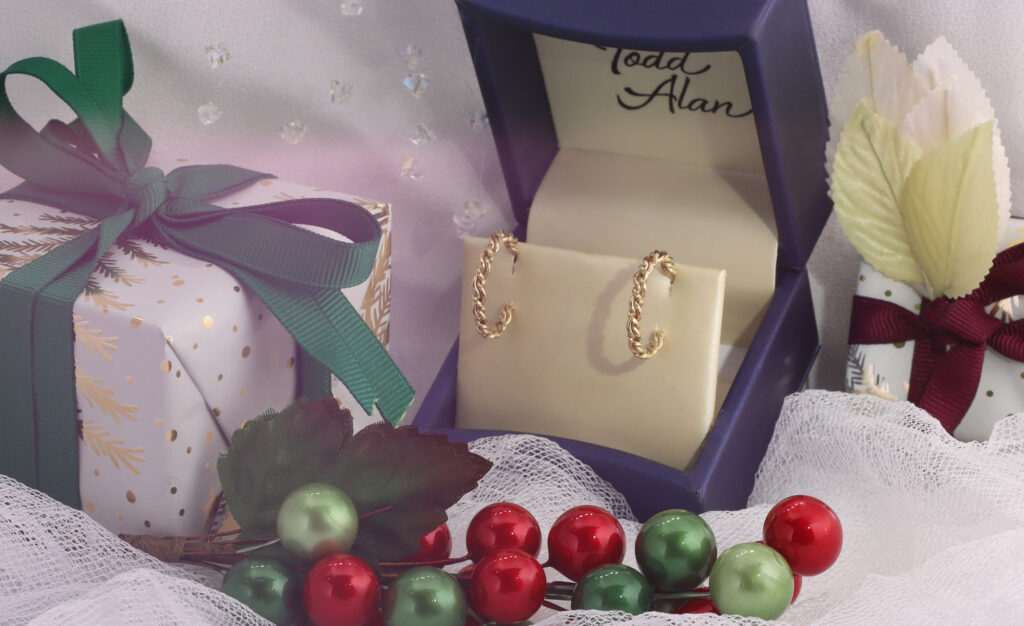 Artistic gold hoop earrings in a blue velvet box, next to a wrapped gift with a green ribbon, amid decorative red and green berries and white lace.