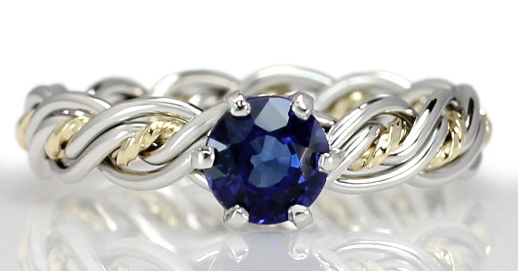 Silver and gold five-strand band ring with a central blue sapphire set in a prong setting, displayed against a reflective white surface.