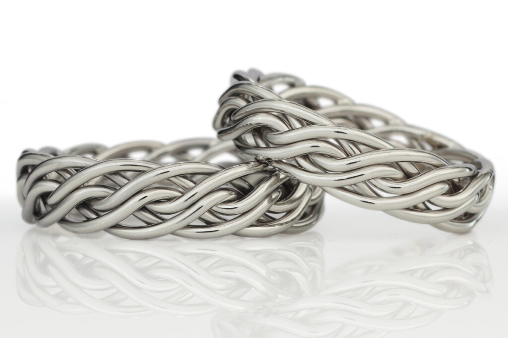 Two intertwined silver bracelets with a detailed, braided design, displayed against a reflective white surface.