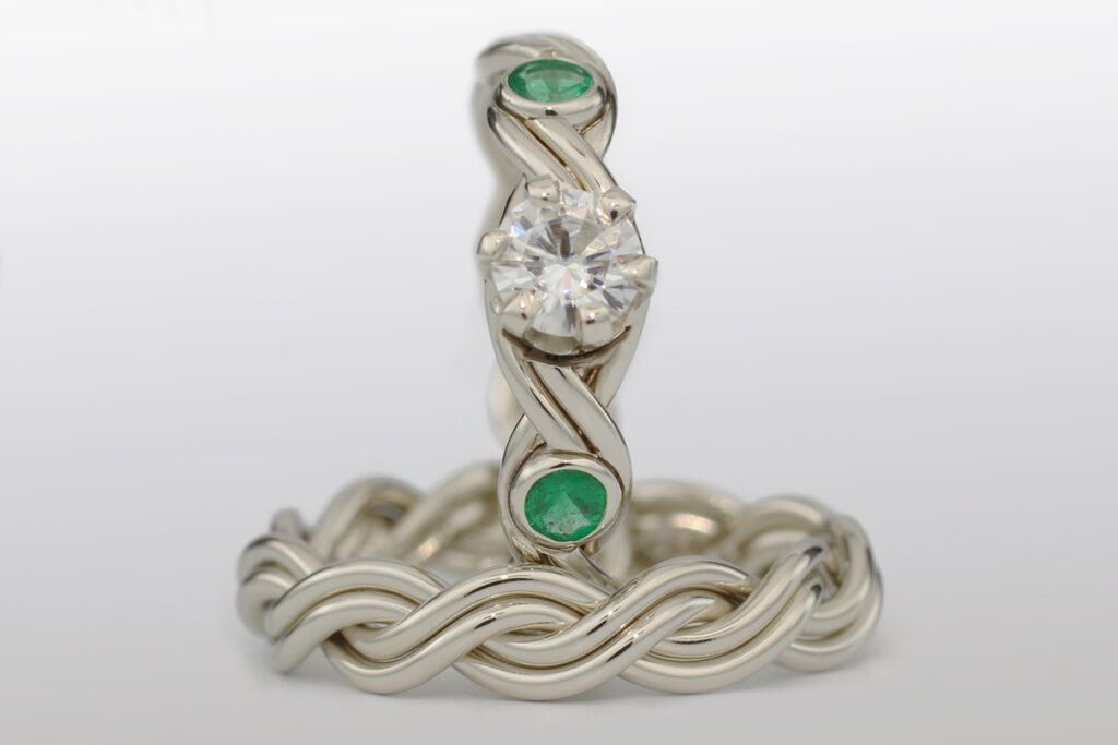 Four strand intertwined silver band ring featuring a central large diamond and two small green gemstones.