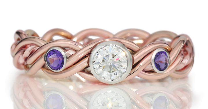 A rose gold wedding ring featuring a central large diamond and two smaller sapphire gemstones, displayed on a reflective surface.
